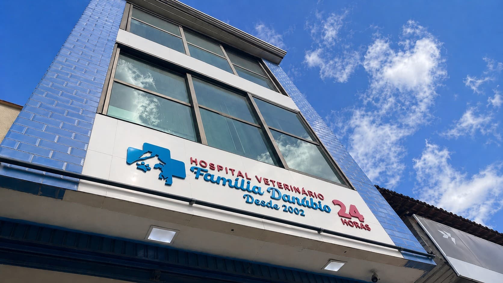Fachada do Hospital Veterinário Família Danúbio com letreiro azul e selo 24 horas, vista de baixo pra cima sob céu azul com nuvens